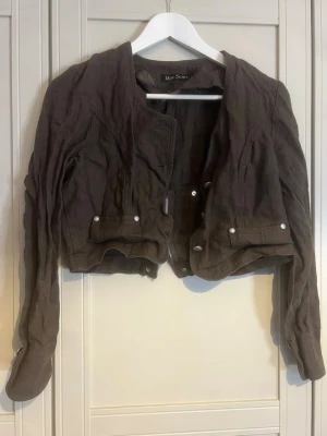 Brun croppad jacka med dragkedja - Snygg brun croppad jacka med långa ärmar och coola detaljer som dragkedja framtill och baktill samt knappar på fickorna. Jackan har en modern, lite loose passform och är tillverkad i ett tyg som ser ut som bomull eller linne.