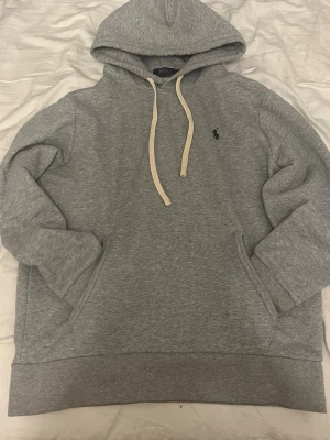 Grå hoodie från Polo Ralph Lauren - Klassisk grå hoodie från Polo Ralph Lauren med vit dragsko och broderad logga på bröstet. Hoodien har känguruficka framtill och ribbade muddar vid ärmar och nederkant. Tillverkad i mjukt sweatshirtmaterial för maximal komfort.
