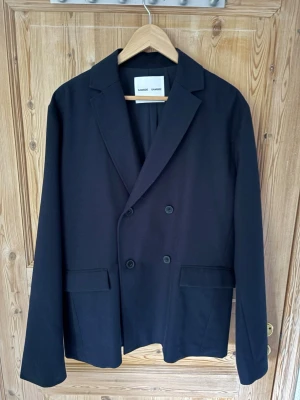 Svart dubbelknäppt kavaj från Samsoe Samsoe - Stilren mörkblå kavaj från Samsoe Samsoe med dubbelknäppning och klassisk krage. Två framfickor med lock och långa ärmar. Passar perfekt till en dressad look och har en tidlös design som funkar till många tillfällen.