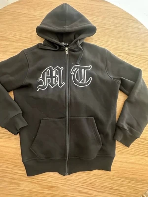 Svart hoodie med tryck och dragkedja - Storlek S. Svart hoodie med dragkedja och huva. På framsidan finns stora bokstäver 'M' och 'T' i vitt tryck, och på ryggen ett stort tryck av en örn. Hoodien har känguruficka och ribbade muddar. Perfekt för en streetwear-look. 