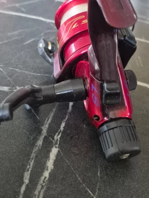 Röd fiskerulle LS-57 - Säljer en röd fiskerulle modell LS-57 med svarta detaljer och guldfärgade markeringar. Handtaget är svart och kroppen är tillverkad i metall. Perfekt till fiskeentusiasten som vill ha en pålitlig rulle till sitt spö.