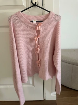 Ljusrosa stickad tröja med rosetter - Supersöt ljusrosa stickad tröja med bred rund halsringning och dekorativa rosetter framtill. Det står att tröjan är storlek 170, men jag skulle snarare säga att de är en stor XS gränsfall mot S 