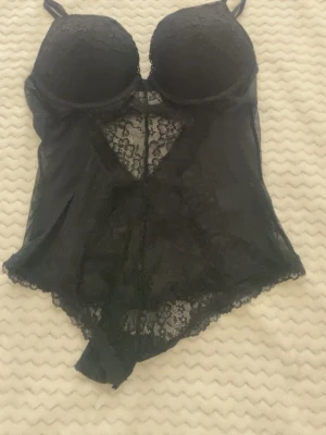 Svart spetsbody med bygel - Snygg svart body i spets med bygelkupor och justerbara axelband. Transparent spets framtill och dekorativt mönster ger en sexig look. 