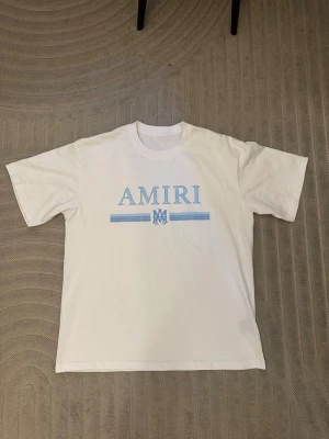 Vit t-shirt från Amiri med blått tryck - Säljer en vit t-shirt från Amiri med ljusblått tryck på bröstet och klassisk rund hals. T-shirten har korta ärmar och är gjord i mjuk bomull. Perfekt för dig som gillar stilrena och trendiga plagg.