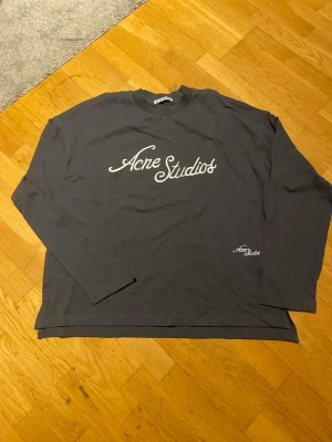 Mörkgrå Acne Studios sleeve - Snygg mörkgrå sleeve från Acne Studios med vit logga tryckt framtill. Tröjan har rund halsringning och långa ärmar. Perfekt för dig som gillar stilrena och trendiga plagg med tydlig branding.