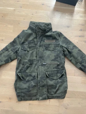 Camo fieldjacket - Säljer en snygg militärinspirerad fieldjacka i grönt camouflagemönster. Jackan har fyra stora fickor framtill, hög krage och dragkedja med vindslå. Tillverkad i slitstark bomullsmix och har en rak passform som ger en cool streetwear-look.