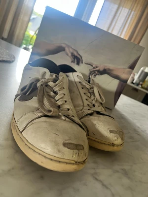 The Gallerist Vita Sneakers - Säljer ett par klassiska vita The Gallerist (Black Heal) sneakers med beige sula och vita snören. Skorna har en enkel och stilren design med svarta detaljer vid hälen. Perfekta för dig som gillar en clean look och vill ha något som funkar till det mesta. Skorna är har en del rejäla defekter, men med rätt lagning samt tvättning kan dem säkert komma till användning igen. Nypris ligger på ca 2700kr, mitt pris 399.