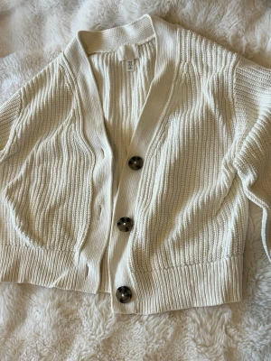 Vit stickad kofta  - Snygg vit stickad kofta från H&M med tre stora bruna knappar framtill. Koftan har v-ringning, ribbade muddar och en croppad passform. Perfekt att slänga över en topp för en chill look.