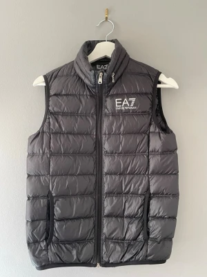 Svart dunväst från EA7 Emporio Armani - Snygg svart dunväst från EA7 Emporio Armani med hög krage och dragkedja framtill. Västen har två fickor med dragkedja och ett stilrent EA7-logotryck på bröstet. Perfekt för lager-på-lager och ger en sportig vibe till din outfit.