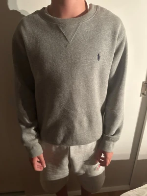 Grå sweatshirt från Polo Ralph Lauren - Klassisk grå sweatshirt från Polo Ralph Lauren med rund halsringning och diskret blå logga broderad på bröstet. Tröjan har ribbade muddar vid ärmslut och nederkant, perfekt för en avslappnad och stilren look.