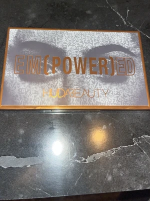 EM(POWER)ED ögonskuggspalett - Snygg ögonskuggspalett från Huda Beauty med namnet EM(POWER)ED. Paletten har ett stilrent fodral i guld och silver med grafiskt ögonmotiv. Perfekt för dig som vill ha många färger samlade i en palett.