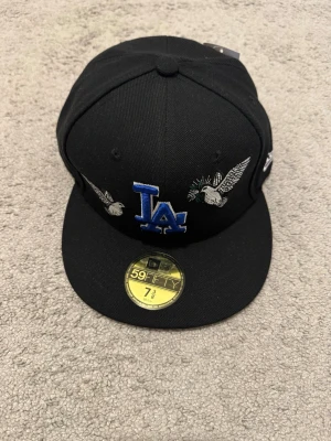 Svart LA Dodgers keps med brodyr - Snygg svart keps från New Era med blå LA Dodgers-logga och broderade duvor på framsidan. Klassisk 59FIFTY-modell med rak skärm och MLB-logga bak. Perfekt för dig som gillar streetwear och sportig stil.