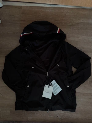 Svart vindjacka från Moncler - Snygg svart vindjacka från Moncler med huva och dragkedja framtill. Jackan har vita och röda detaljer på huvan samt två fickor med tryckknappar. Tillverkad i ett lätt och tåligt material, perfekt till hösten! NFC-märket funkar