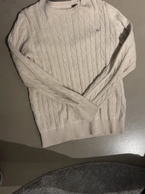Beige kabelstickad tröja från GANT - Säljer en beige kabelstickad tröja från GANT i premium bomull. Tröjan har rund halsringning, ribbade muddar och diskret GANT-logga på bröstet. Perfekt för dig som gillar klassisk och stilren design.
