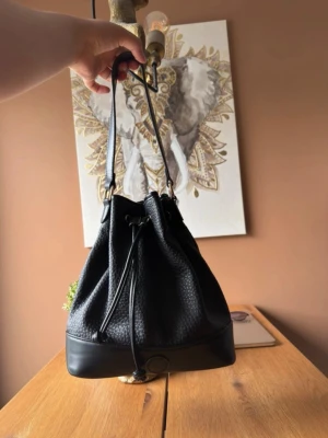Svart bucket bag i skinnimitation - Snygg svart bucket bag med dragskostängning och axelrem. Väskan är gjord i skinnimitation med struktur och har detaljer i metall. Botten är förstärkt och slät, vilket ger en cool kontrast till resten av väskan. Perfekt för dig som gillar stilrena accessoarer.