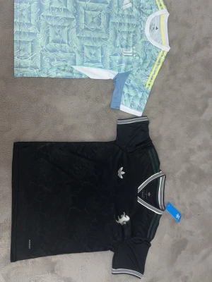 Adidas Juventus och svart och blå T-shirt  1 för 250kr 2 för 550kr - Två snygga sporttröjor från Adidas. Ena är en Juventus-tröja i ljusblått, vitt och gult med grafiskt mönster och klubbmärke. Den andra är en svart retroinspirerad tröja med vita detaljer, Adidas-logga och lejonmärke. Perfekta för fotboll eller streetwear.