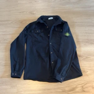 Svart OverShirt - Stilren svart skjorta med knappstängning och två bröstfickor. På vänster ärm sitter en rund patch med gul och grön logga. Skjortan har klassisk krage och långa ärmar med manschettknappar. Perfekt för en trendig och avslappnad look.