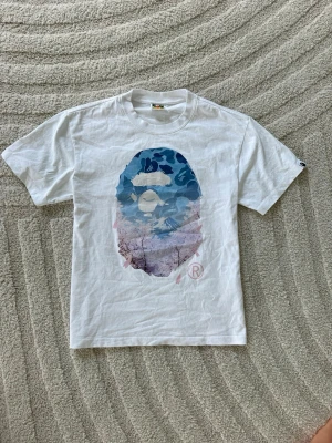 Vit BAPE t-shirt med blått tryck - Säljer en vit t-shirt från BAPE med ikoniskt blått camo-tryck på bröstet. T-shirten har rund hals och korta ärmar. Materialet är mjuk bomull och passformen är normal. Perfekt för dig som gillar streetwear och vill sticka ut lite extra.