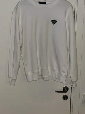 Prada Vit sweatshirt med logga på bröstet - Snygg vit sweatshirt med rund hals och ribbade muddar. Tröjan har en liten svart logga på vänstra bröstet och är tillverkad i mjukt material, perfekt för en clean och stilren look. Passar till allt och är enkel att matcha med jeans eller sweatpants.