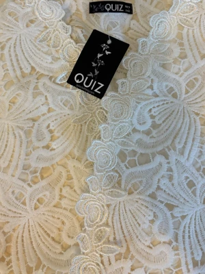 Vit spetsklänning från QUIZ - Elegant vit klänning från QUIZ med vackert blommigt spetsmönster och dekorativa broderade detaljer längs mitten. Klänningen har en romantisk känsla och är perfekt för speciella tillfällen. Materialet är spets med fodrad insida för extra komfort.