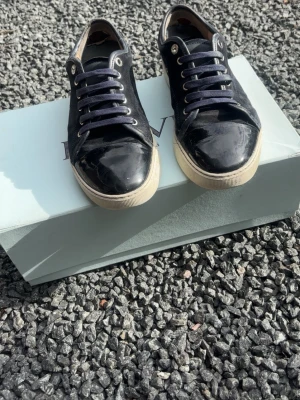 Lanvin dbb1  - Säljer ett par svarta sneakers med coola lackdetaljer på tån och klassiska vita sulor. Skorna har svarta snören och en stilren design som passar till det mesta. Materialmix av textil och lack ger en snygg kontrast och modern känsla.