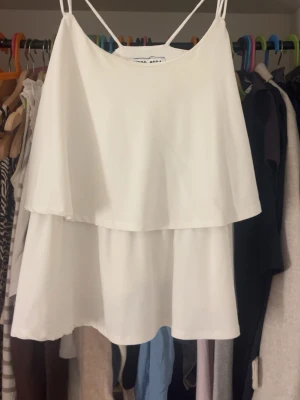 Vit volang tröja från Vero Moda - Säljer en vit kort tröja från Vero Moda med tunna axelband och dubbla lager tyg för en luftig känsla. Tröjan har en enkel och stilren design, perfekt för varma dagar. Materialet känns lätt och bekvämt.