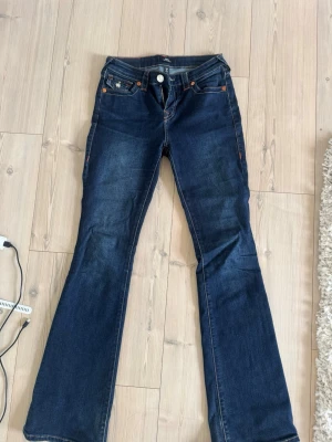 Mörkblå bootcut jeans med kontrastsömmar från true religion - Säljer ett par mörkblå bootcut jeans från true religion med snygga kontrastsömmar och dekorativa fickor bak. Jeansen har klassisk femficksdesign och utsvängda ben. Perfekta för dig som gillar retrovibbar och vill ha ett par jeans som sticker ut lite extra. Passar mig som är 172, men även till kortare personer 