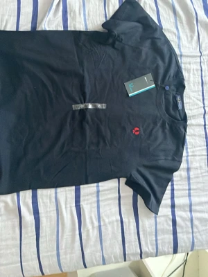 Svart t-shirt från Fred Perry - Snygg svart t-shirt från Fred Perry med klassisk rund hals och röd broderad logga på bröstet. Tillverkad i mjuk bomull för skön känsla och enkel stil. Perfekt till jeans eller shorts för en clean look.