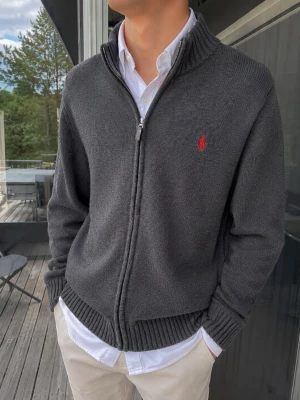 Polo Ralph Lauren Full Zip – Grå, Storlek M - Stilren Polo Ralph Lauren full zip-tröja i mörkgrå nyans med klassisk röd hästlogga broderad på bröstet. Stickad struktur med ribbade muddar och krage samt praktisk dragkedja framtill. Perfekt att bära över en vit skjorta för en klassisk, preppy och tidlös look. Storlek M. Tröjan är i mycket fint skick 