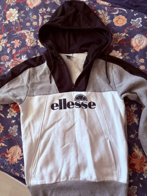 Ellesse hoodie i grått, vitt och svart - Snygg hoodie från Ellesse i grått, vitt och svart med stor logga på bröstet. Tröjan har dragkedja, huva med snören och ribbade muddar vid ärmsluten. Perfekt för en avslappnad och sportig stil. Tillverkad i mjukt sweatshirtmaterial.
