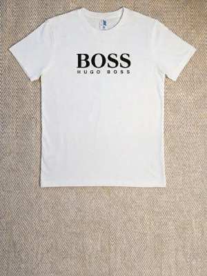 Vit t-shirt Hugo Boss - Säljer en stilren vit t-shirt från Hugo Boss med klassisk svart logga på bröstet. T-shirten har rund halsringning och korta ärmar, perfekt för en clean och enkel look. Tillverkad i mjuk bomull för skön komfort.