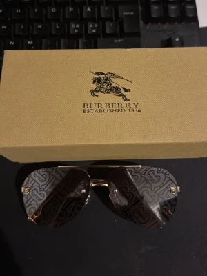 Burberry aviator solglasögon guld - Säljer ett par snygga Burberry solglasögon i aviator-modell med guldfärgade bågar och tonade glas med Burberry-monogram. Kommer med originaletui i klassiskt rutigt mönster och putsduk. Perfekt accessoar för att lyfta din stil.