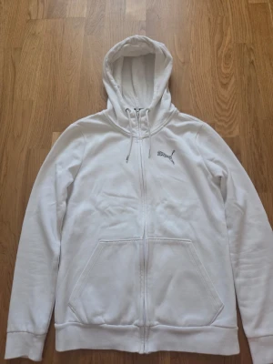 Vit zip hoodie från Puma - Säljer en vit hoodie från Puma med dragkedja framtill och två stora fickor. Hoodien har justerbar huva med snören och Puma-logga broderad på bröstet. Tillverkad i mjukt sweatshirtmaterial, perfekt för en avslappnad stil.