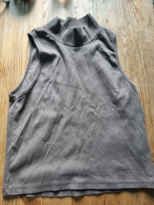 Grå ribbad ärmlös polotopp - Säljer en stilren grå polotopp i ribbad bomull. Toppen är ärmlös och har en hög krage som ger en trendig look. Perfekt att matcha med jeans eller kjol för en enkel men snygg outfit.