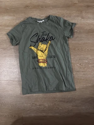 Grön t-shirt med shaka-tryck - Snygg grön t-shirt med ett stort gult shaka-tryck och texten 'The Shaka' samt 'Baja California' på framsidan. T-shirten har korta ärmar och en avslappnad passform. Perfekt för dig som gillar surfvibbar och coola prints.
