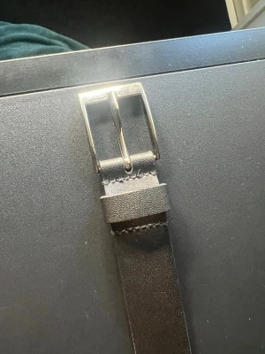 Svart klassiskt skärp i äkta läder - Snyggt och stilrent svart bälte i läder med silverfärgat metallspänne. Perfekt till jeans eller kostymbyxor för en clean look. Enkel design utan mönster och med justerbara hål för bästa passform.