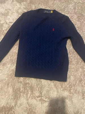 Mörkblå kabelstickad tröja Polo Ralph Lauren - Kabelstickad mörkblå tröja från Polo Ralph Lauren med rund halsringning och den klassiska röda logotypen broderad på bröstet. Tröjan har långa ärmar och ribbade muddar vid ärmslut och nederkant. Perfekt för en stilren look.