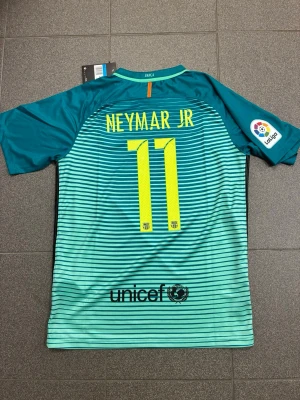 Barcelona Neymar Jr tröja  - Snygg Barcelona matchtröja i turkos med svarta ränder, Neymar Jr och nummer 11 på ryggen. Tröjan har korta ärmar, La Liga-märke på ärmen och Qatar Airways-logga framtill. Perfekt för fotbollsfans som vill sticka ut på planen eller läktaren. Turkos aktig färg 