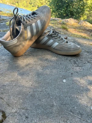 Grå Adidas sneakers i mocka - Säljer ett par grå Adidas sneakers i mocka med klassiska vita ränder på sidorna och gummisula. Skorna har lågt skaft och snörning framtill. Perfekta för dig som gillar stilren och sportig look.