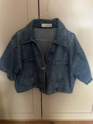 Kortärmad croppad jeansjacka blå - Snygg croppad jeansjacka i klassisk blå denim med korta ärmar och två stora bröstfickor. Jackan har en avslappnad passform, bred krage och knäppning framtill med metallknappar. Perfekt för en trendig och avslappnad look. Storlek XL men passar mig som har S 