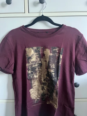 Vinröd t-shirt med guldtryck - Säljer en vinröd t-shirt med korta ärmar och ett stort abstrakt tryck i guld och svart på framsidan. T-shirten har en klassisk rund halsringning och är gjord i mjuk bomull. Perfekt för dig som vill ha något unikt och stilrent i garderoben.