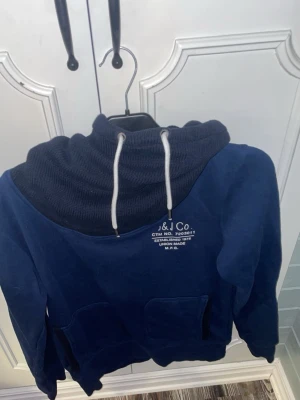 Mörkblå hoodie med tryck och huva - Säljer en mörkblå hoodie med stor, ribbad huva och vita dragsnören. Tryck på bröstet med texten 'J&J Co.'