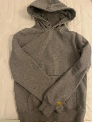 Grå hoodie från Carhartt - Säljer en stilren grå hoodie från Carhartt med klassisk huva och magficka. Hoodien har ribbade muddar och en liten gul Carhartt-logga broderad vid ärmslutet. Perfekt för en avslappnad och trendig look.