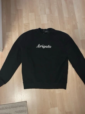 Svart sweatshirt med Arigato-tryck - Säljer en svart sweatshirt med vit text 'Arigato' på bröstet. Tröjan har rund halsringning, långa ärmar och ribbade muddar vid ärmslut och nederkant. Perfekt för en clean och stilren look. Normalstor i storleken (S). Använd men i bra skick.