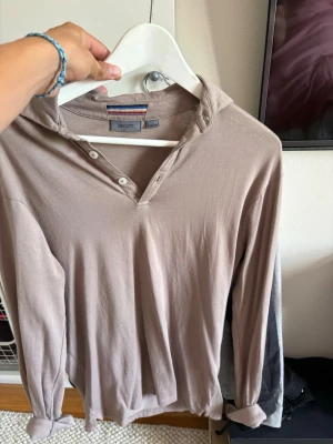 Beige långärmad tröja från Snoot - Långärmad beige tröja från Snoot med knappar vid halsen och avslappnad passform. Tillverkad i mjukt material och har en klassisk krage. Perfekt för en chill och stilren look.