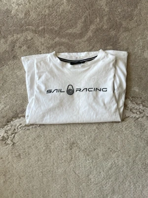 Vit Sail Racing t-shirt med tryck - Vit t-shirt från Sail Racing med svart logotyptryck på bröstet. Klassisk rund hals och korta ärmar. Perfekt för en avslappnad och sportig stil. Tillverkad i mjuk bomull för skön komfort.