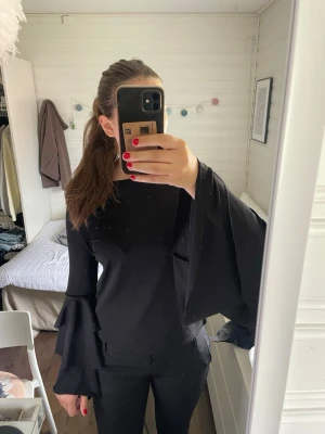 Svart topp med volangärmar - Säljer en svart topp med rund hals och långa, vida ärmar med dubbla volangdetaljer. Har små defekter som syns på bild. Perfekt för dig som vill ha något extra till din outfit.