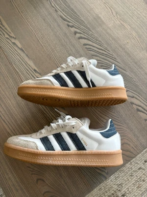 Adidas Samba OG vita sneakers - Klassiska Adidas Samba OG sneakers i vitt läder med beige mockadetaljer och svarta ränder på sidorna. Skorna har en brun gummisula och Adidas-logga på plösen. Perfekta för en stilren och sportig look.