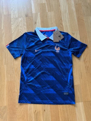 Blå Frankrike fotbollströja Nike - Säljer en blå fotbollströja från Nike med Frankrikes landslagsmärke och två stjärnor på bröstet. Tröjan har ett mönster i olika blå nyanser, vit krage och korta ärmar. Perfekt för dig som älskar fotboll och vill stötta Les Bleus.