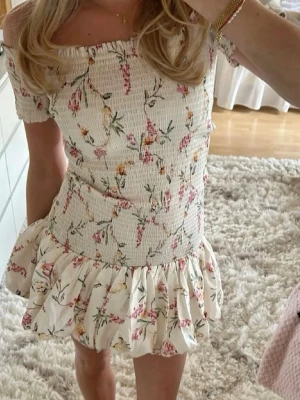 Blommig offshoulder kortklänning - Supersöt kortklänning med offshoulder och volangkant. Klänningen är vit med blommigt mönster i rosa, gult och grönt. Smockad överdel och luftig kjol ger en feminin och somrig look. Perfekt för varma dagar och picknickar.